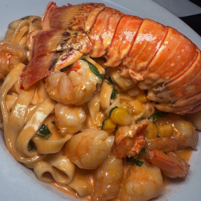Lobster tail and shrimp pasta with creamy sauce, corn, and greens on a white plate.